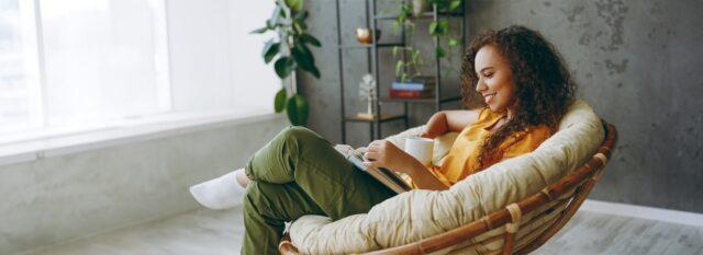 Woman relaxing at home in a round wicker chair with a book and a hot drink
